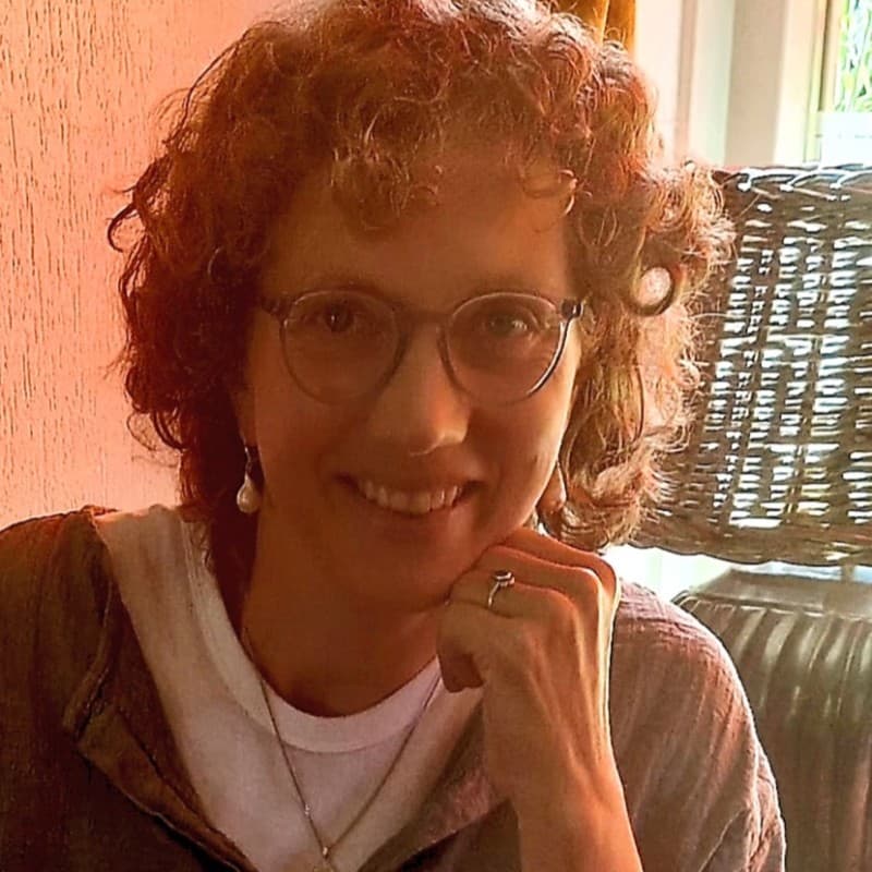 Smiling woman with curly reddish-brown hair and glasses, resting her chin on her hand.