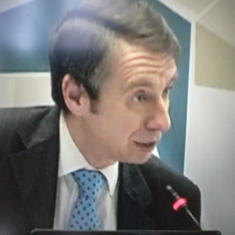 Man in a dark suit and blue polka-dot tie speaking into a microphone.