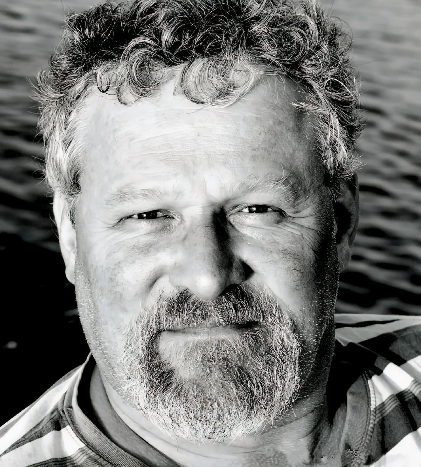 Black and white portrait of a bearded man with curly hair against rippling water.