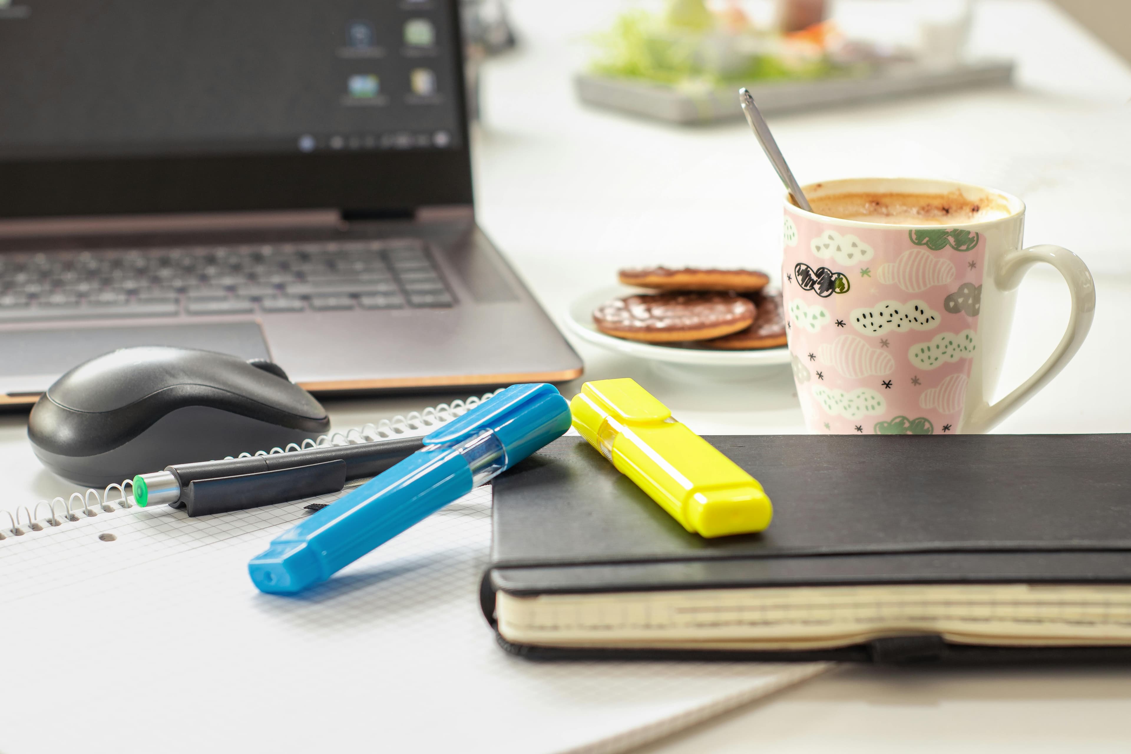 Workspace featuring a laptop, notebooks, highlighters, a patterned coffee mug, and chocolate-covered cookies.
